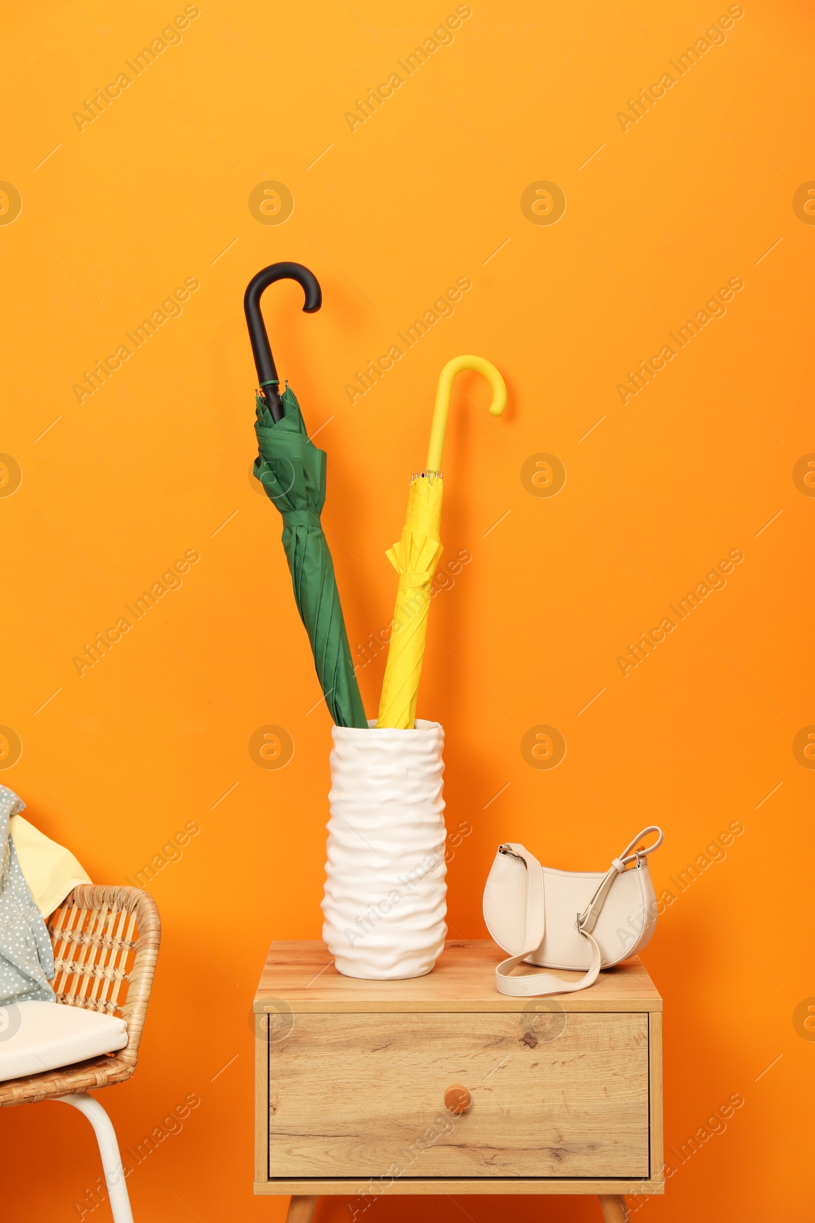 Photo of Colorful umbrellas in holder and bag on wooden side table near orange wall indoors