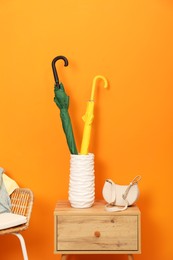 Photo of Colorful umbrellas in holder and bag on wooden side table near orange wall indoors