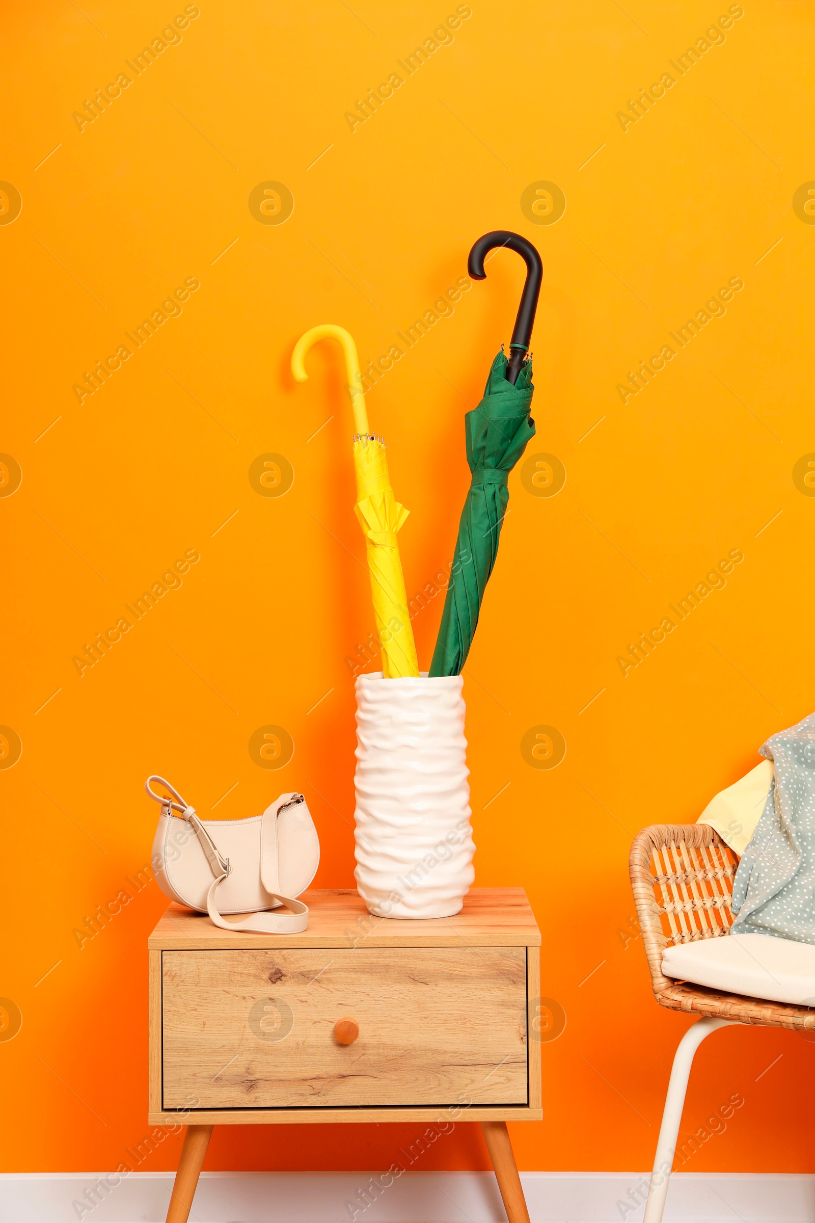 Photo of Colorful umbrellas in holder and bag on wooden side table near orange wall indoors