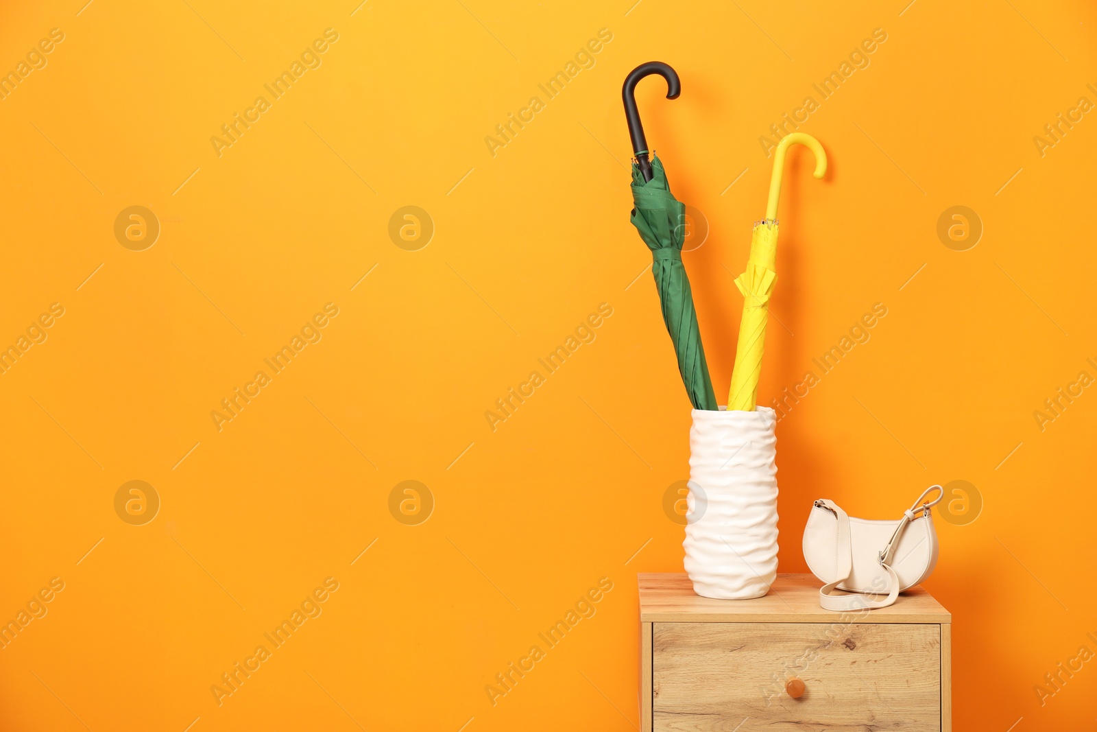 Photo of Colorful umbrellas in holder and bag on side table near orange wall indoors, space for text