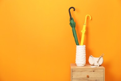 Photo of Colorful umbrellas in holder and bag on side table near orange wall indoors, space for text