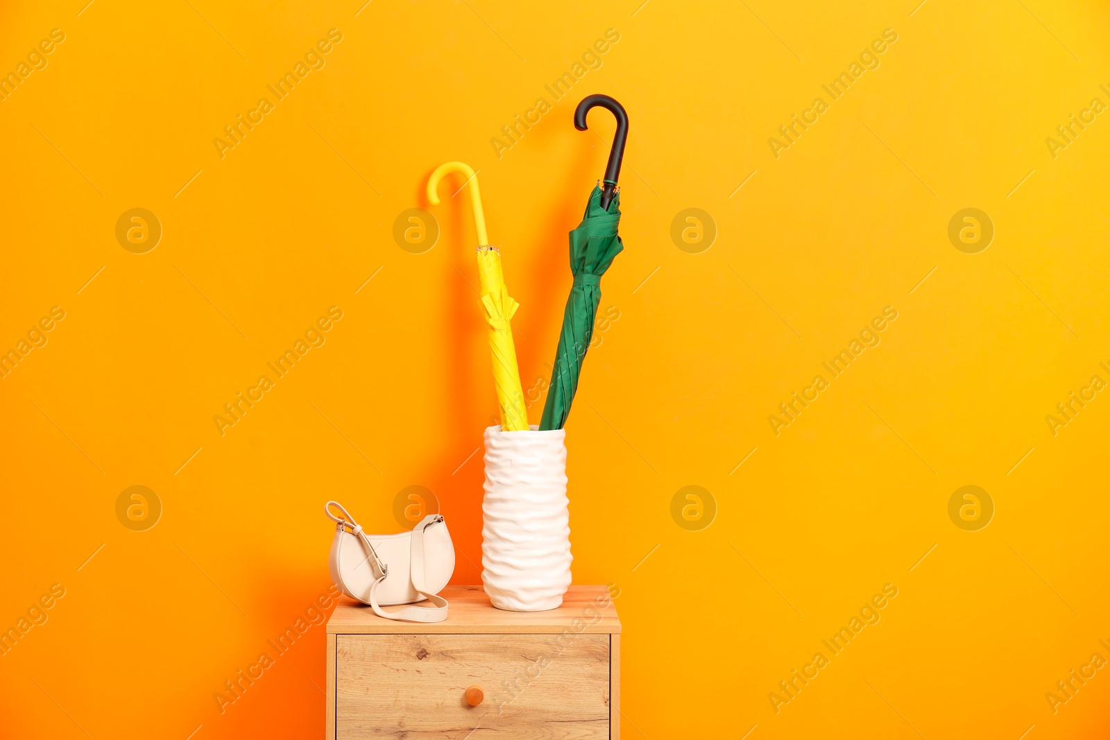 Photo of Colorful umbrellas in holder and bag on wooden side table near orange wall indoors