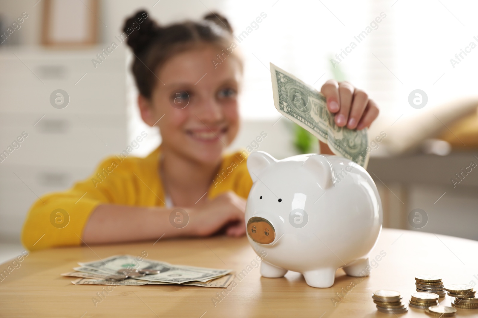 Photo of Pocket money. Cute girl putting dollar banknote into piggy bank at wooden table indoors, selective focus