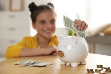 Pocket money. Cute girl putting dollar banknote into piggy bank at wooden table indoors, selective focus