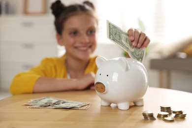 Pocket money. Cute girl putting dollar banknote into piggy bank at wooden table indoors, selective focus