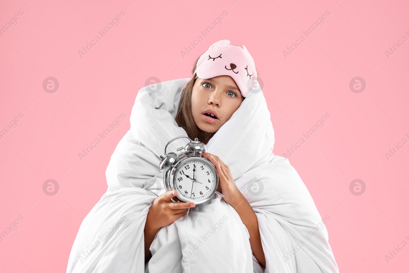 Photo of Overslept girl with sleep mask and alarm clock wrapped in blanket on pink background