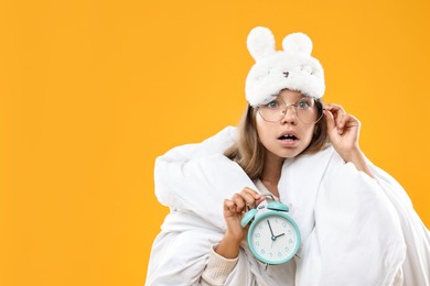 Overslept girl with sleep mask and alarm clock wrapped in blanket on orange background, space for text