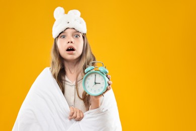 Photo of Overslept girl with sleep mask and alarm clock wrapped in blanket on orange background, space for text