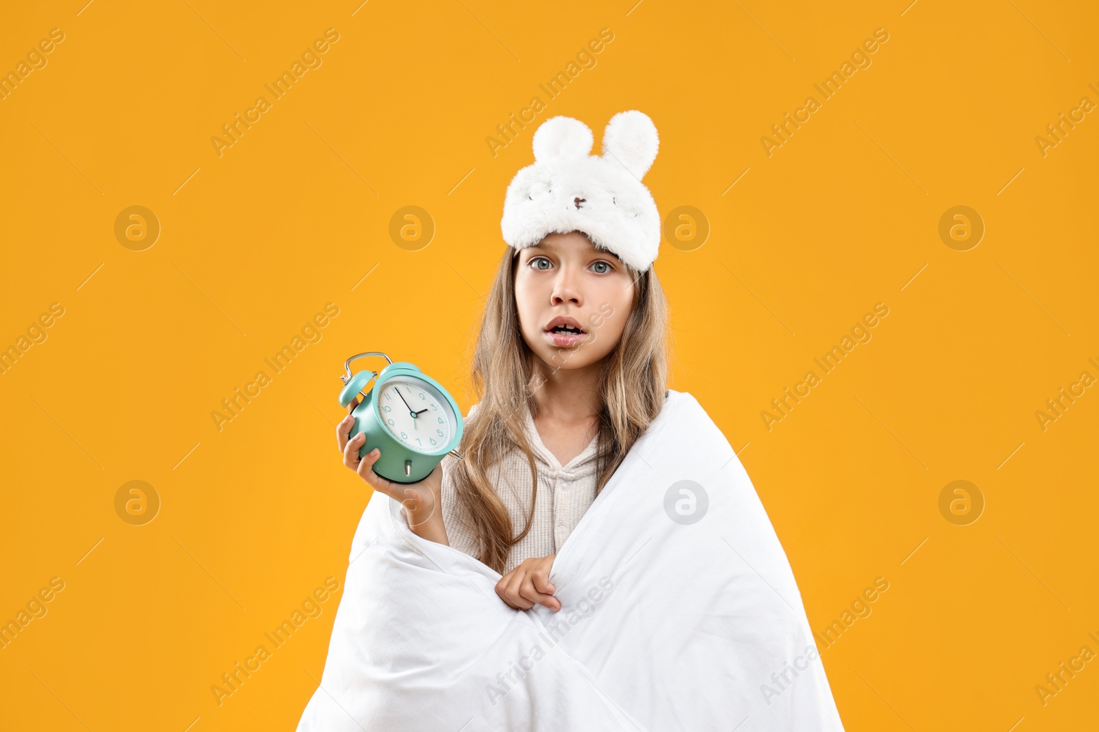 Photo of Overslept girl with sleep mask and alarm clock wrapped in blanket on orange background