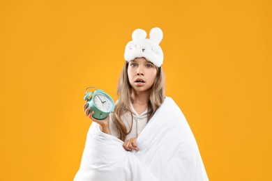 Photo of Overslept girl with sleep mask and alarm clock wrapped in blanket on orange background