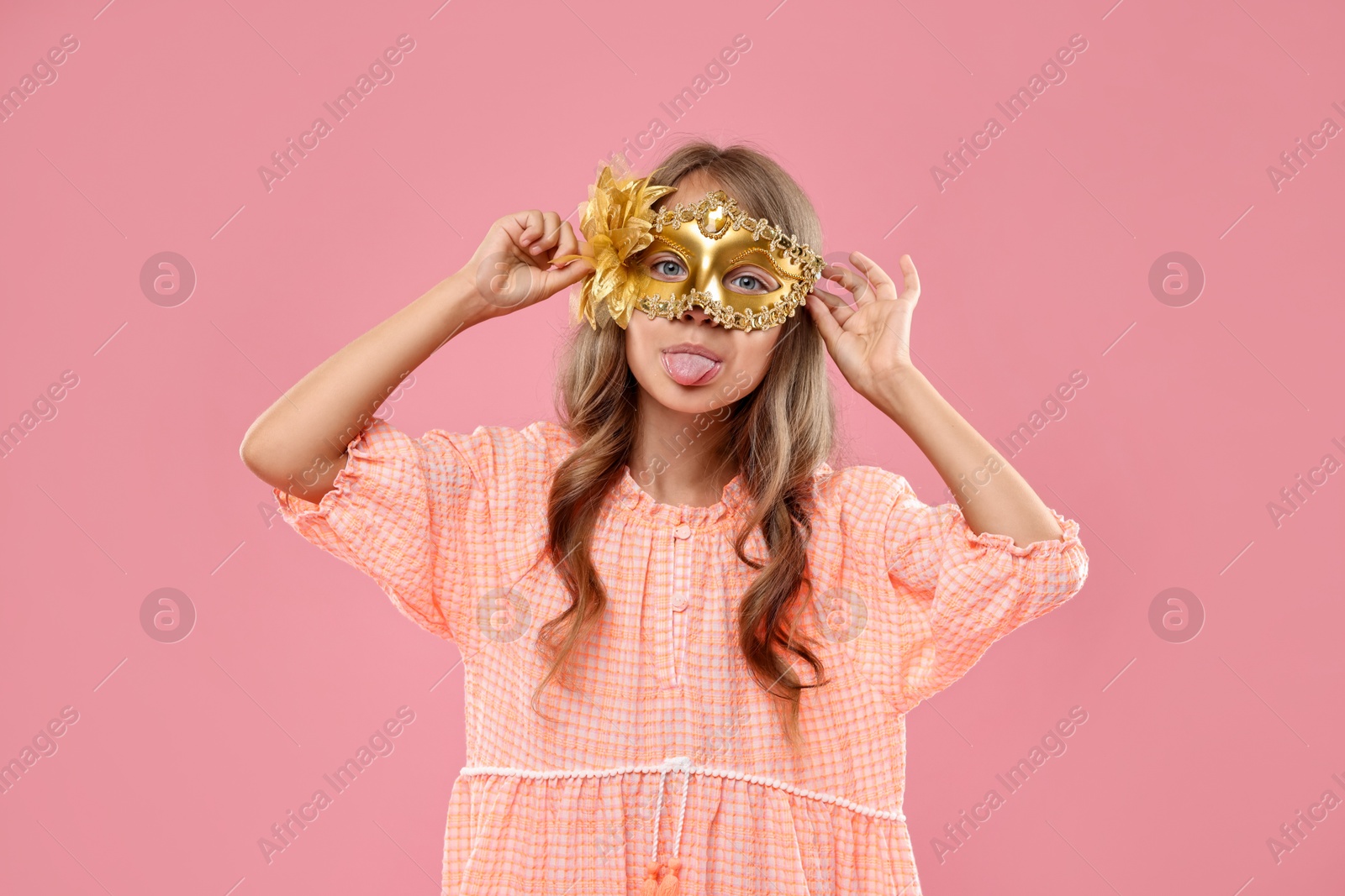 Photo of Cute girl wearing carnival mask and showing tongue on pink background