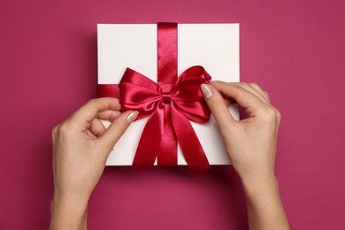 Photo of Woman decorating gift box with bow on dark rose background, top view