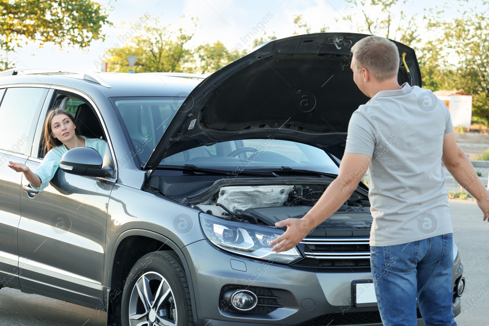 Photo of Stressed man standing near broken car while woman sitting in auto outdoors