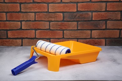 Photo of Paint roller and yellow tray on light textured table against red brick wall