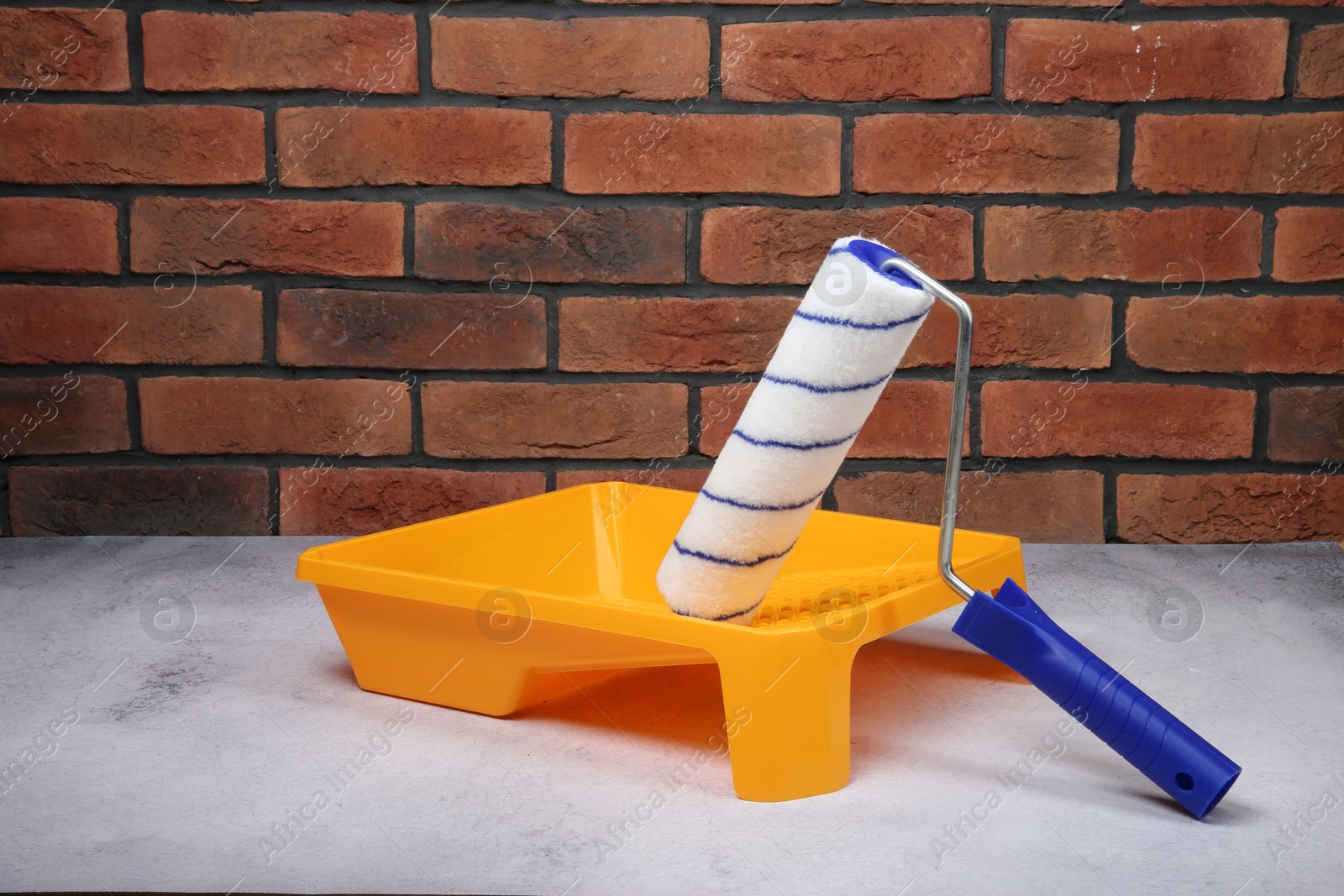 Photo of Paint roller and yellow tray on light textured table against red brick wall