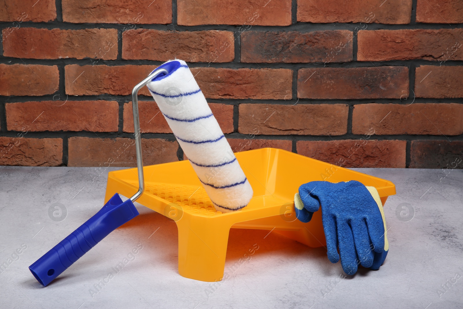 Photo of Paint roller, gloves and yellow tray on light textured table against red brick wall