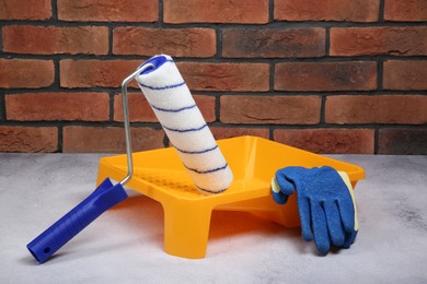 Photo of Paint roller, gloves and yellow tray on light textured table against red brick wall