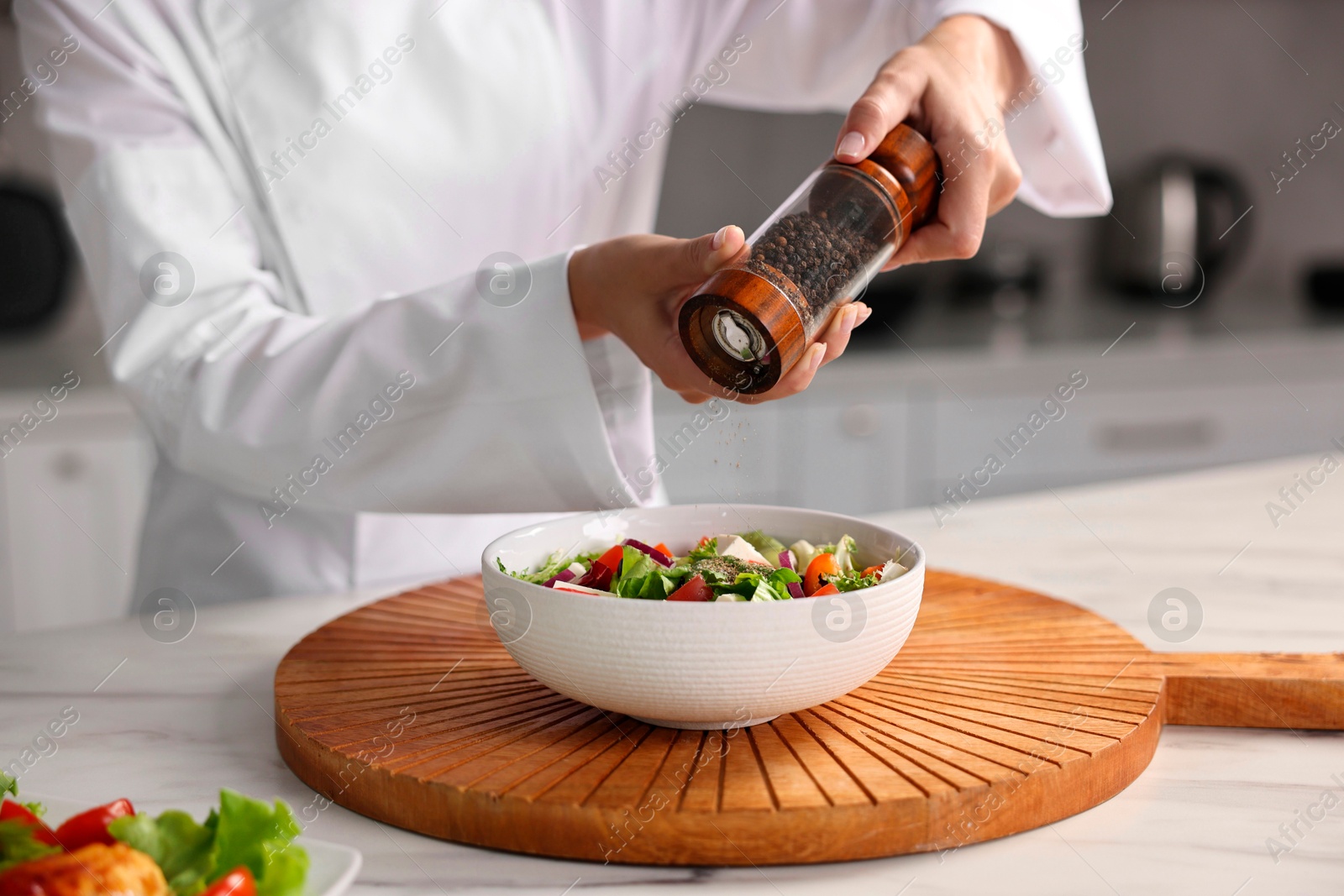 Photo of Professional chef adding pepper to delicious salad at white marble table in kitchen, closeup