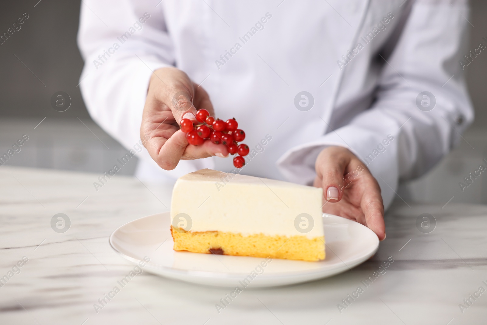 Photo of Professional chef decorating delicious cheesecake with red currants at white marble table in kitchen, closeup