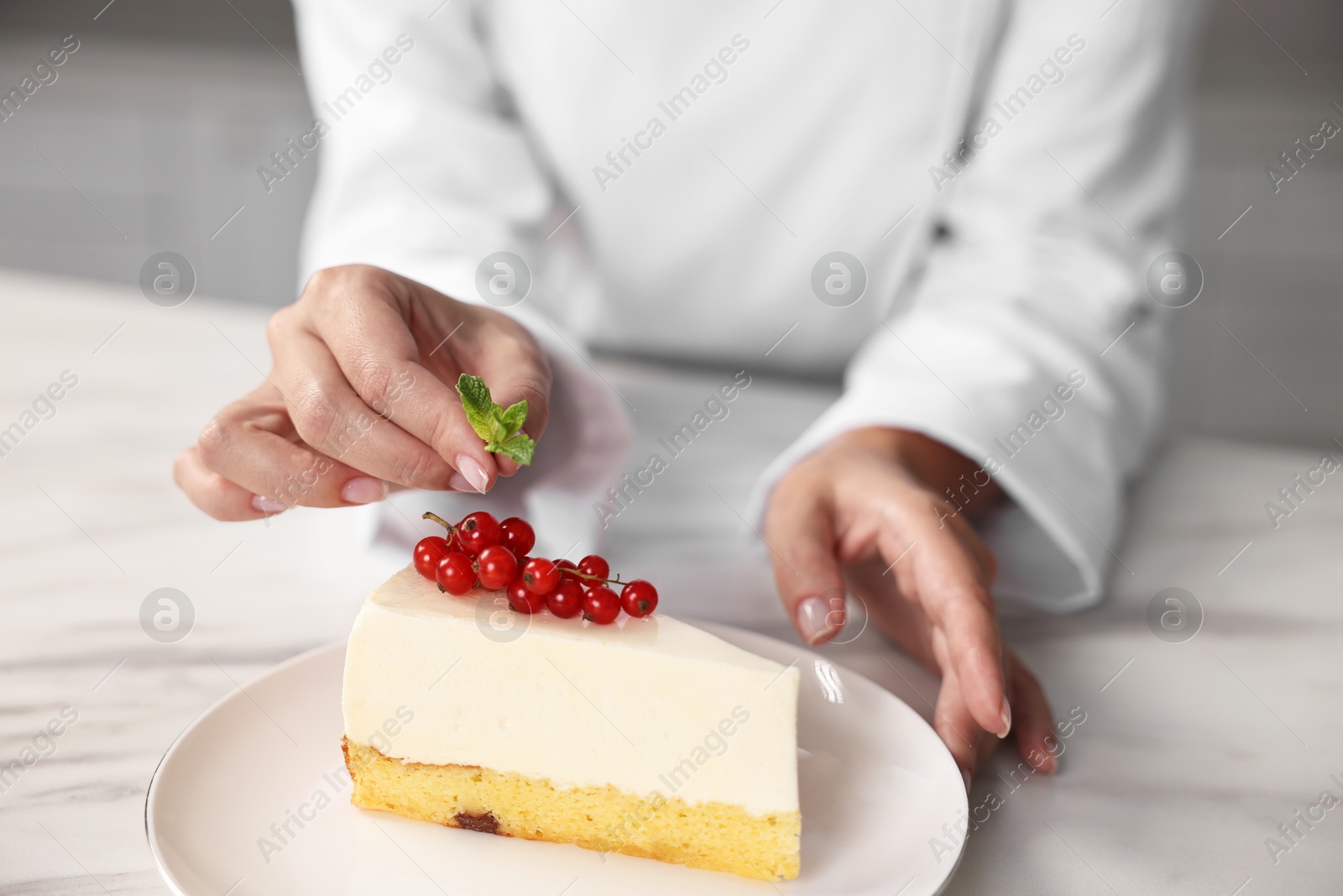 Photo of Professional chef decorating delicious cheesecake with red currants at white marble table in kitchen, closeup