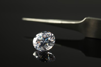 Photo of Beautiful shiny diamond and tweezers on black mirror surface, closeup