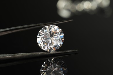 Photo of Tweezers with beautiful shiny diamond on black mirror surface, closeup