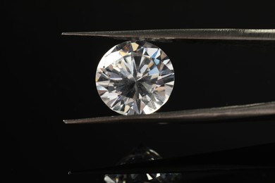 Photo of Tweezers with beautiful shiny diamond on black mirror surface, closeup