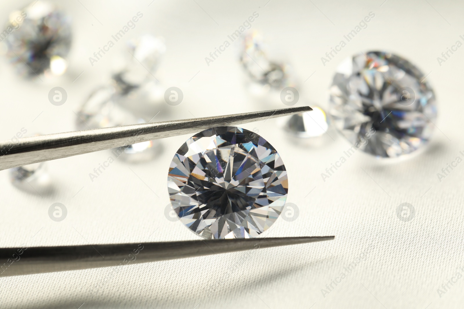 Photo of Tweezers with beautiful shiny diamonds on white fabric, closeup