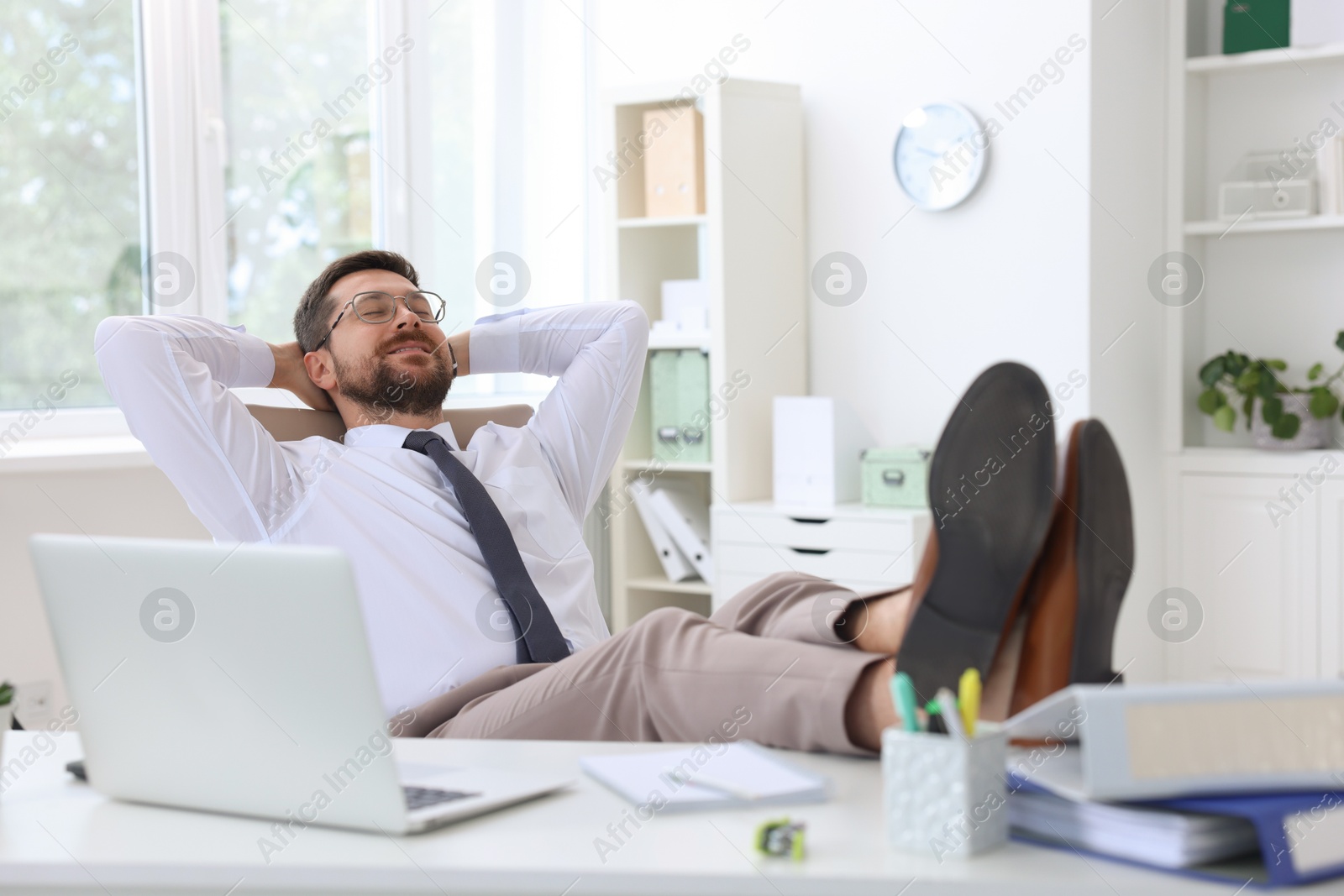 Photo of Smiling businessman with hands behind his head holding legs on table in office. Break time