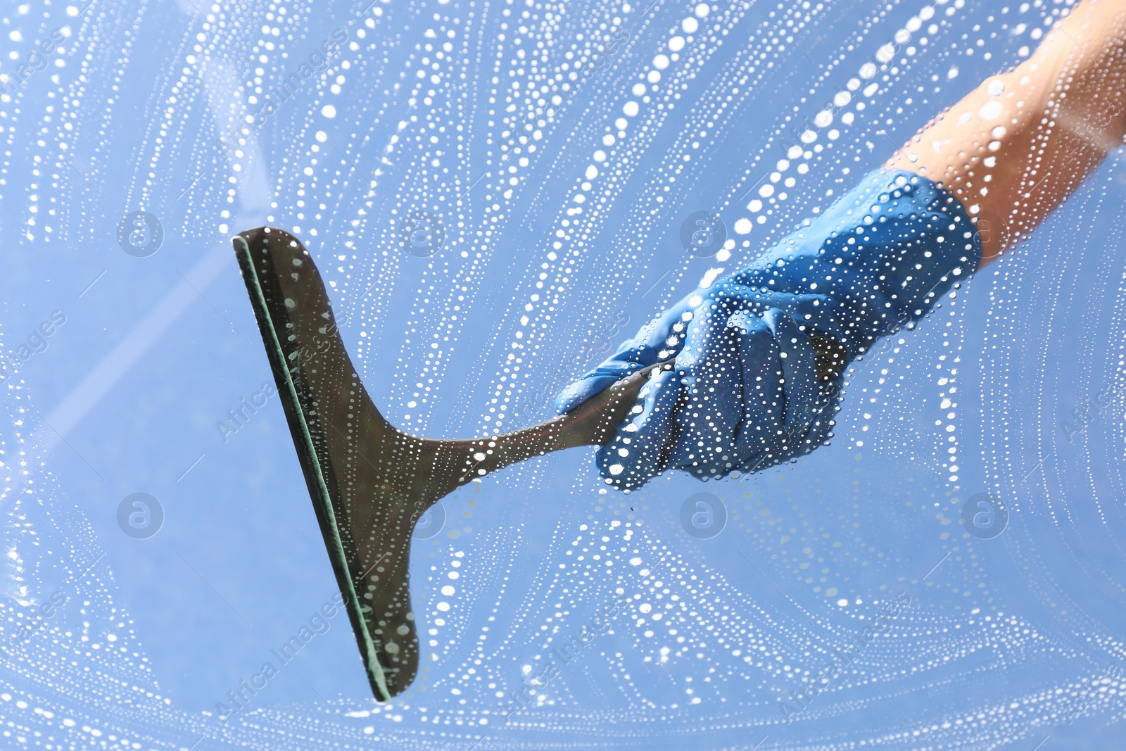 Photo of Woman washing window with squeegee tool against blue sky, closeup