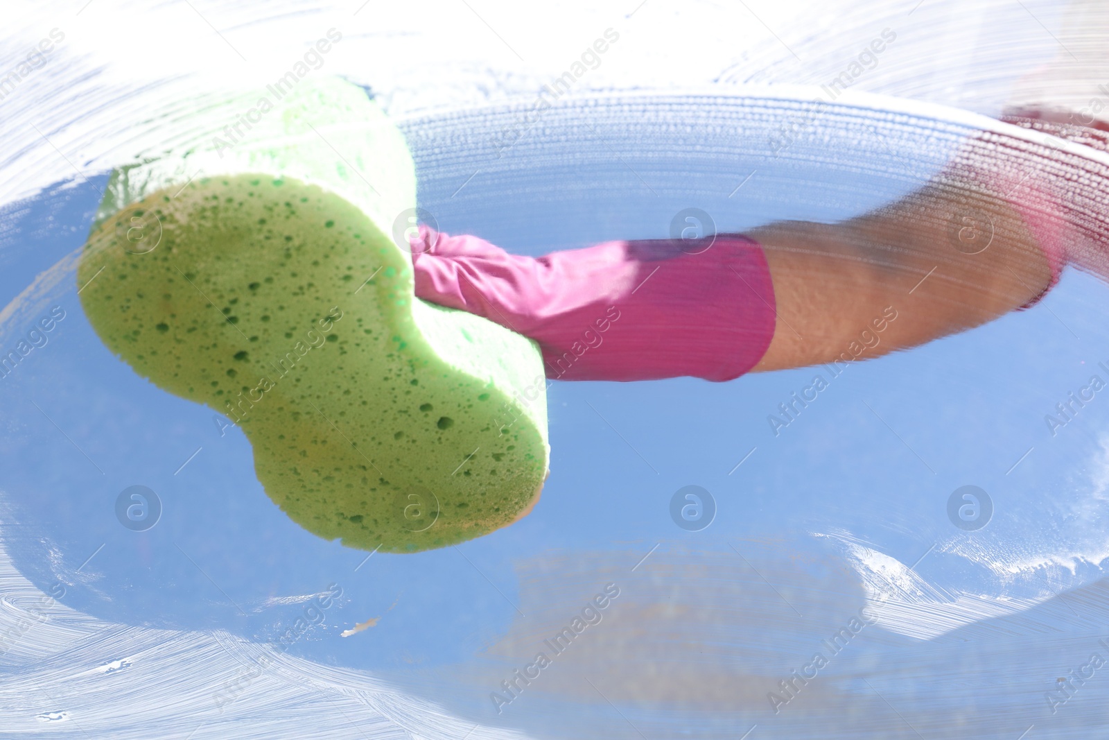 Photo of Woman washing window with sponge against blue sky, closeup