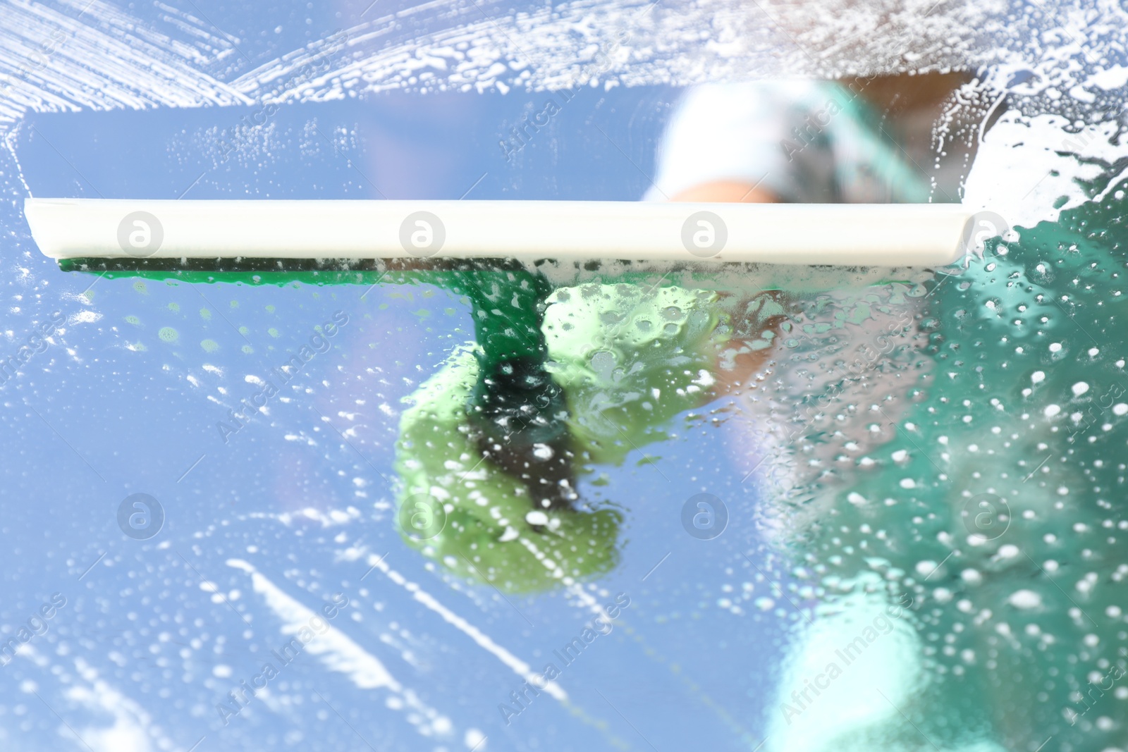 Photo of Woman washing window with squeegee tool against blue sky, closeup