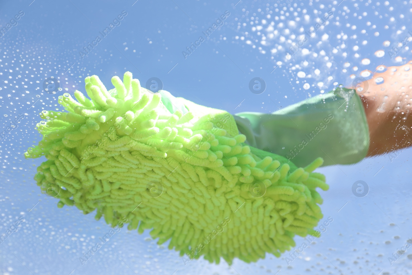 Photo of Woman washing window with microfiber mitt against blue sky, closeup