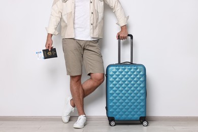 Man with suitcase, passport and ticket near light wall indoors, closeup