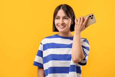 Beautiful woman with smartphone listening to voice message on yellow background