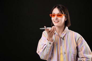 Photo of Beautiful woman in sunglasses recording voice message via smartphone on black background, space for text