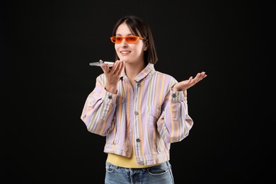 Photo of Beautiful woman in sunglasses recording voice message via smartphone on black background