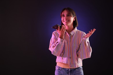 Beautiful woman recording voice message via smartphone on dark background with neon lights, space for text