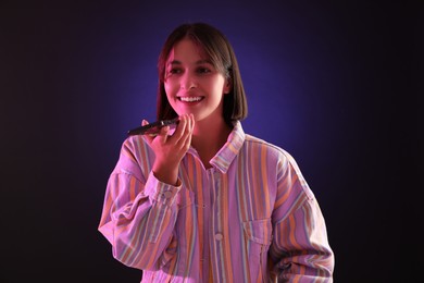 Photo of Beautiful woman recording voice message via smartphone on dark background with neon lights