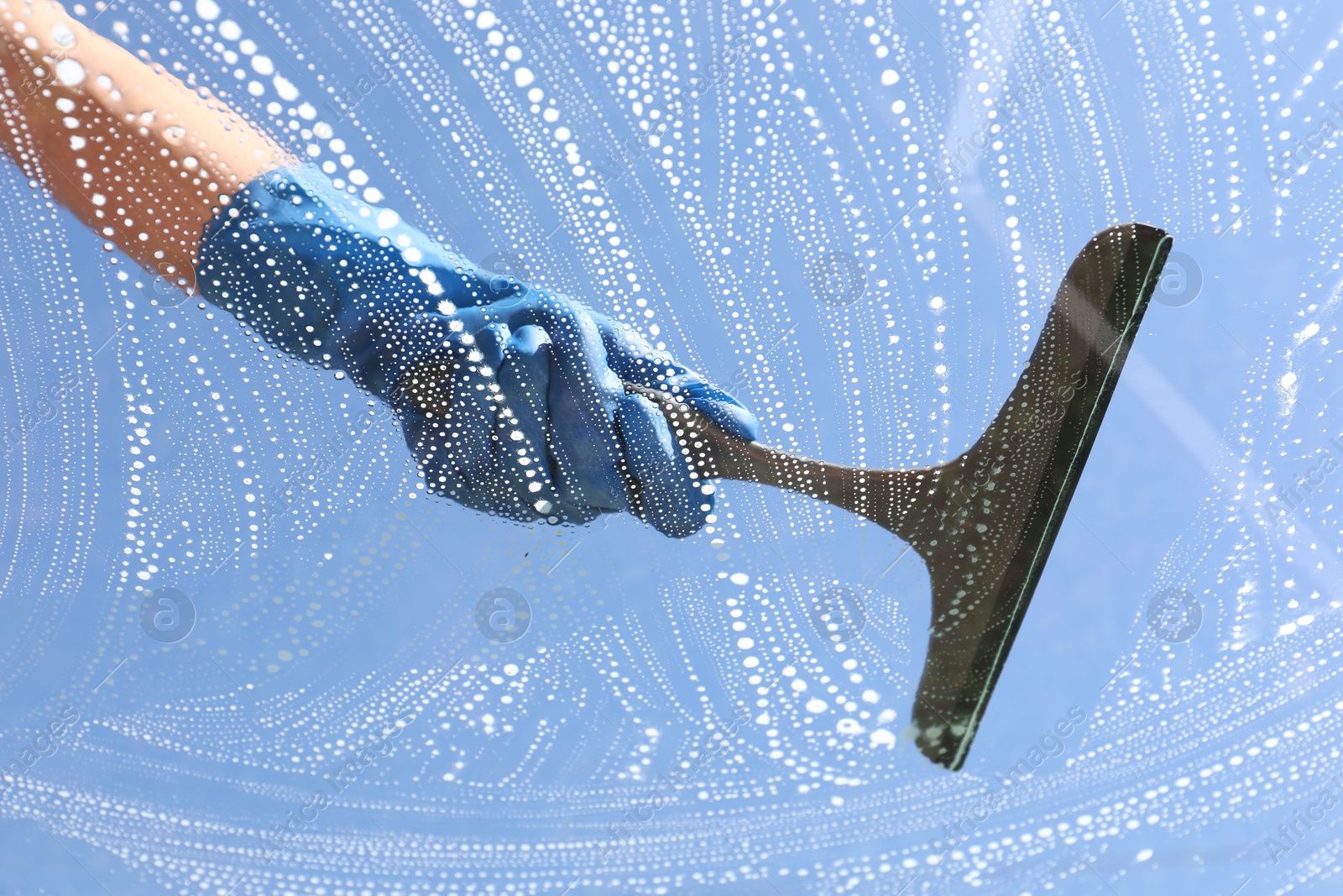 Photo of Woman washing window with squeegee tool against blue sky, closeup