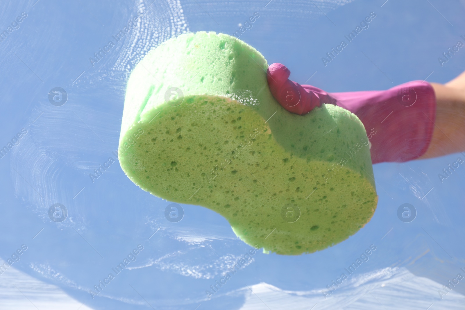 Photo of Woman washing window with sponge against blue sky, closeup