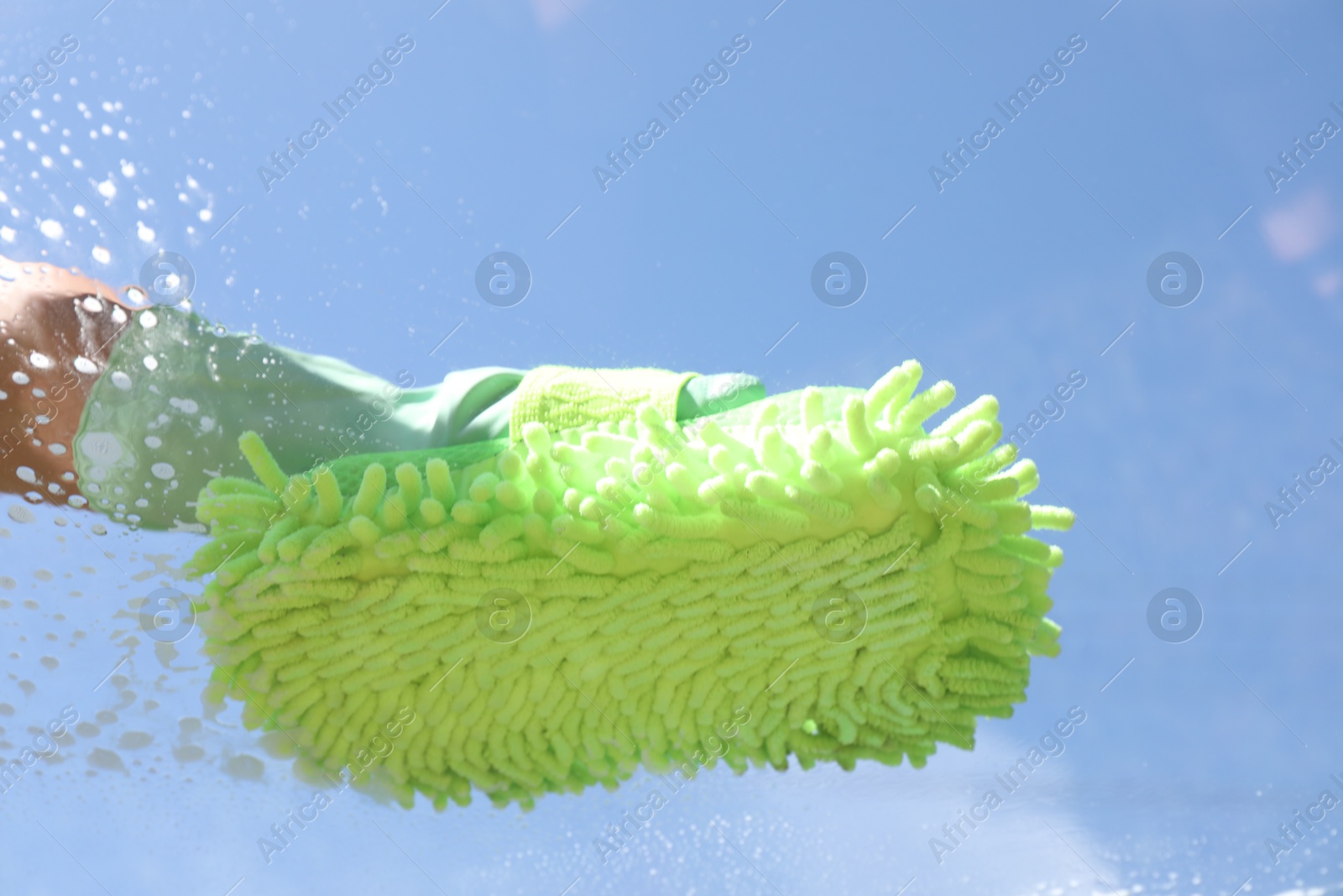 Photo of Woman washing window with microfiber mitt against blue sky, closeup