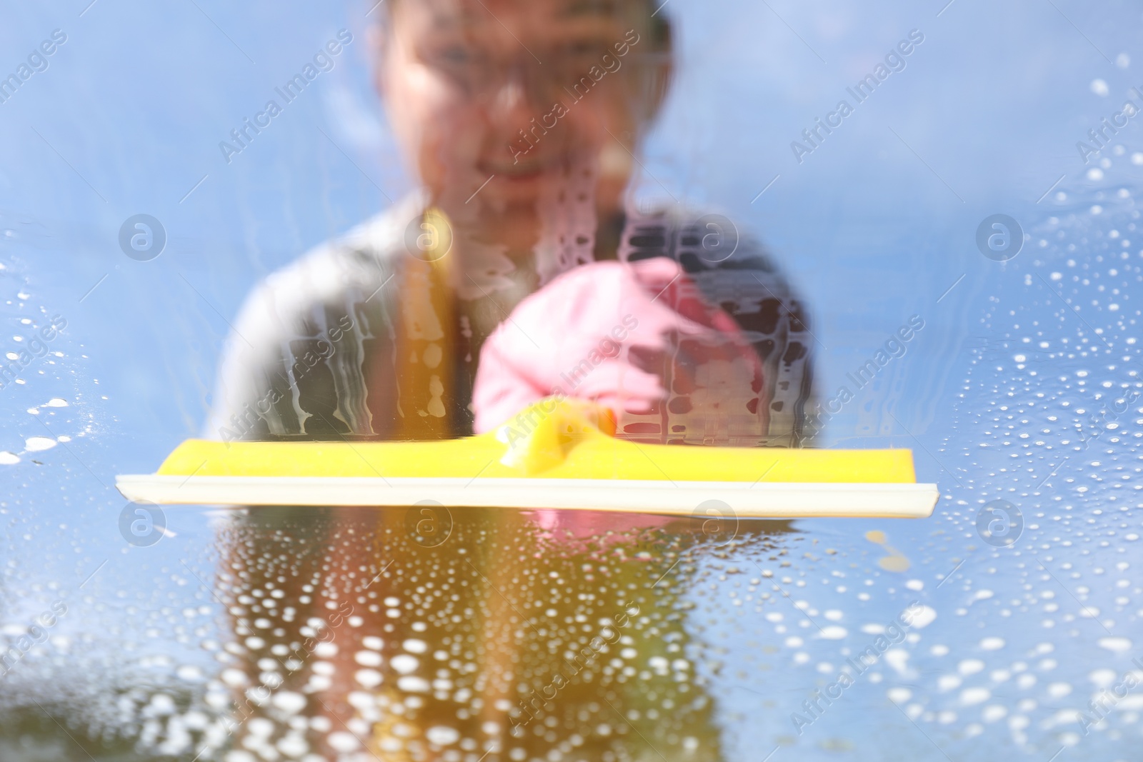 Photo of Woman washing window with squeegee tool against blue sky, closeup