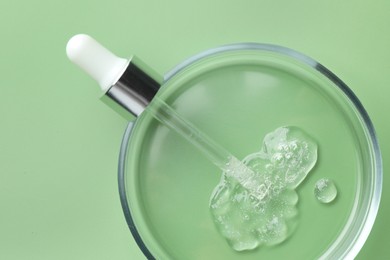 Photo of Petri dish with cosmetic product and pipette on green background, top view