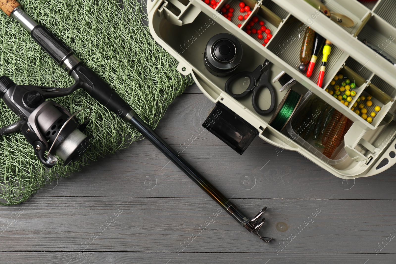 Photo of Different fishing tackles in box, rod with spinning reel and net on grey wooden table, top view