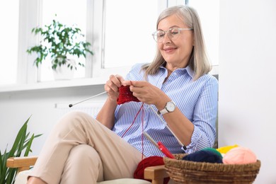 Smiling senior woman knitting on armchair at home, low angle view