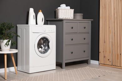 Photo of Washing machine, detergents, chest of drawers and houseplant on table indoors