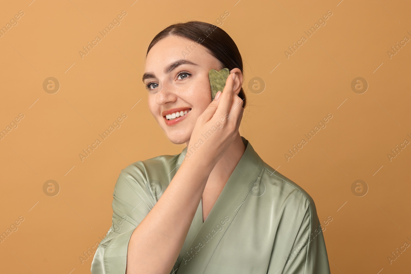 Photo of Beautiful young woman doing facial massage with gua sha tool on beige background