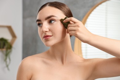 Photo of Beautiful young woman doing facial massage with gua sha tool indoors