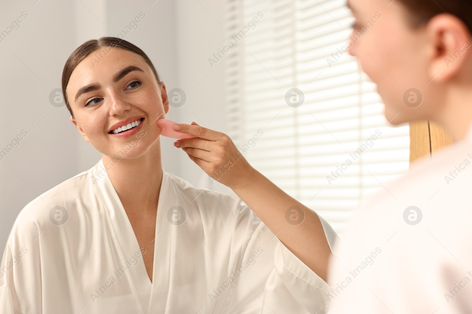 Photo of Beautiful young woman doing facial massage with gua sha tool near mirror at home